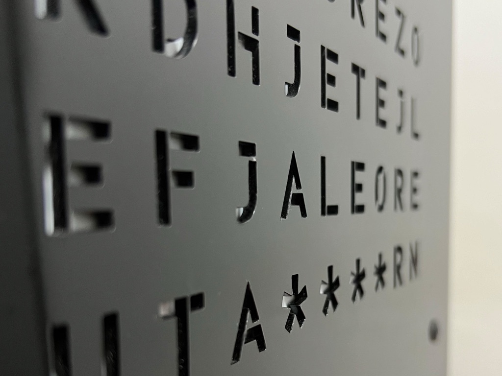 Fotografi makro e hapjeve të shkronjave të prera me lazer dhe yjeve të minutave në pllakën e FjaleOre